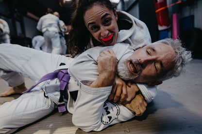 A man and a woman wearing white BJJ gis branded with GMA. Woman has a gift wrap off the man’s arms and man is exaggerating the tightness of the hold. 